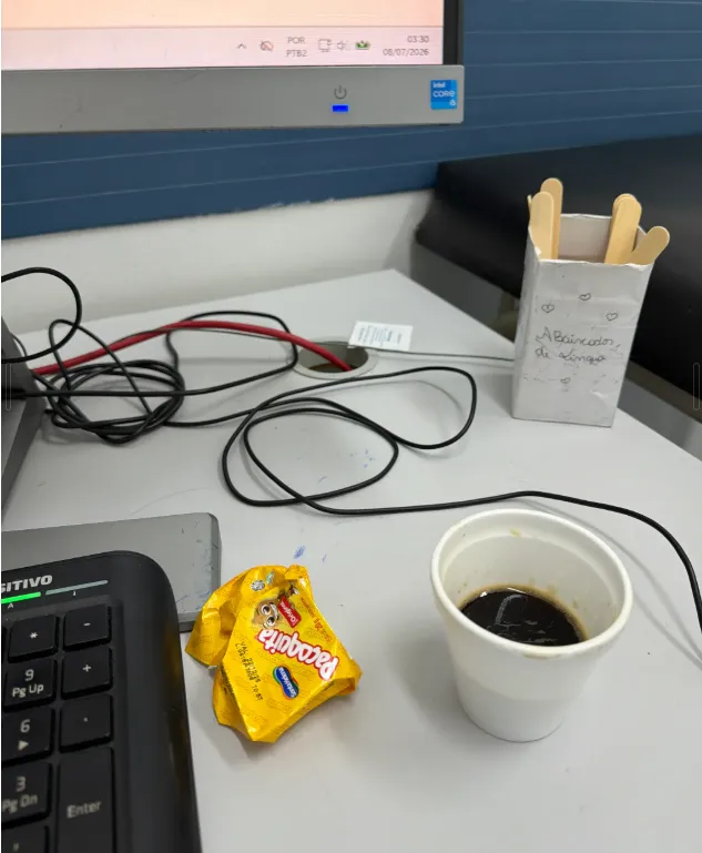 Mesa de trabalho com teclado, embalagem de paçoca aberta e copo de café, com o relógio da tela marcando 03:30 de 08/07/2026